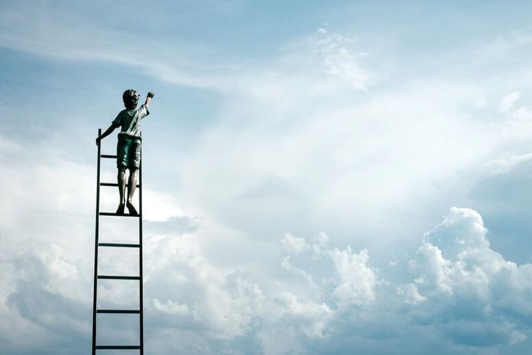 Goal setting is a lot like this young boy reaching an arm up while standing near the top of a ladder in the clouds