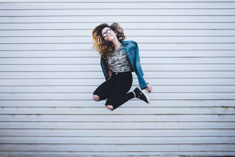 Young woman joyfully jumping up in front of a white concrete wall reacts to the power of positive thinking