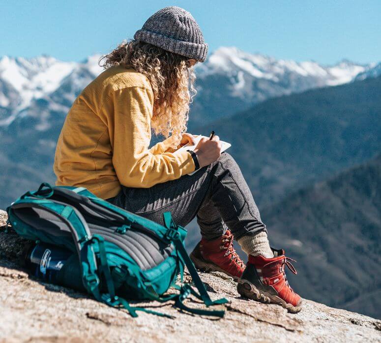Young woman wearing hiking gear sits on rock facing mountains to do some journal writing